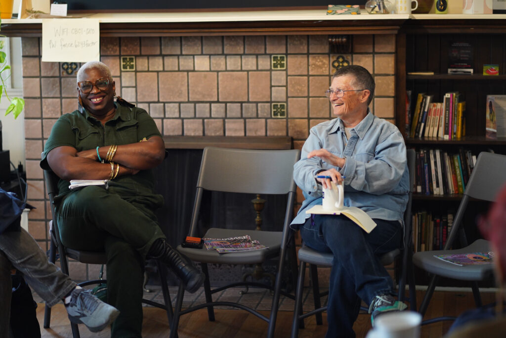 NPN's Director of Racial Justice and Movement Building Sage Crump and Co-founder of the James and Grace Lee Boggs Center Shea Howell are captured smiling during a visit to the Boggs Center. They are seated in front of a fireplace with a bookshelf behind them. There are copies of Riverwise, a community-based magazine covering local activism in Detroit, on the chairs beside Crump and Howell.