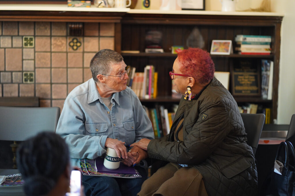Co-founder of the James and Grace Lee Boggs Center Shea Howell and NPN Board Member and Founder of Diaspora Vibe Cultural Arts Incubator (DVCAI) Rosie Gordon-Wallace are captured in conversation during a visit to the Boggs Center. They are seated in front of a bookshelf and fireplace. Gordon-Wallace is tenderly gripping Howell's hands as they speak. 