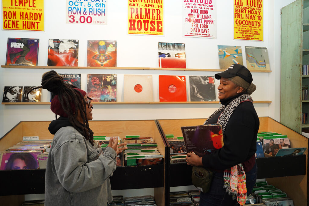 Two attendees chat in front of record bins at NPN's Community Reception at Spot Lite Detroit. The person on the right is holding a record in their hands while the person on the left is gesticulating with their hands.