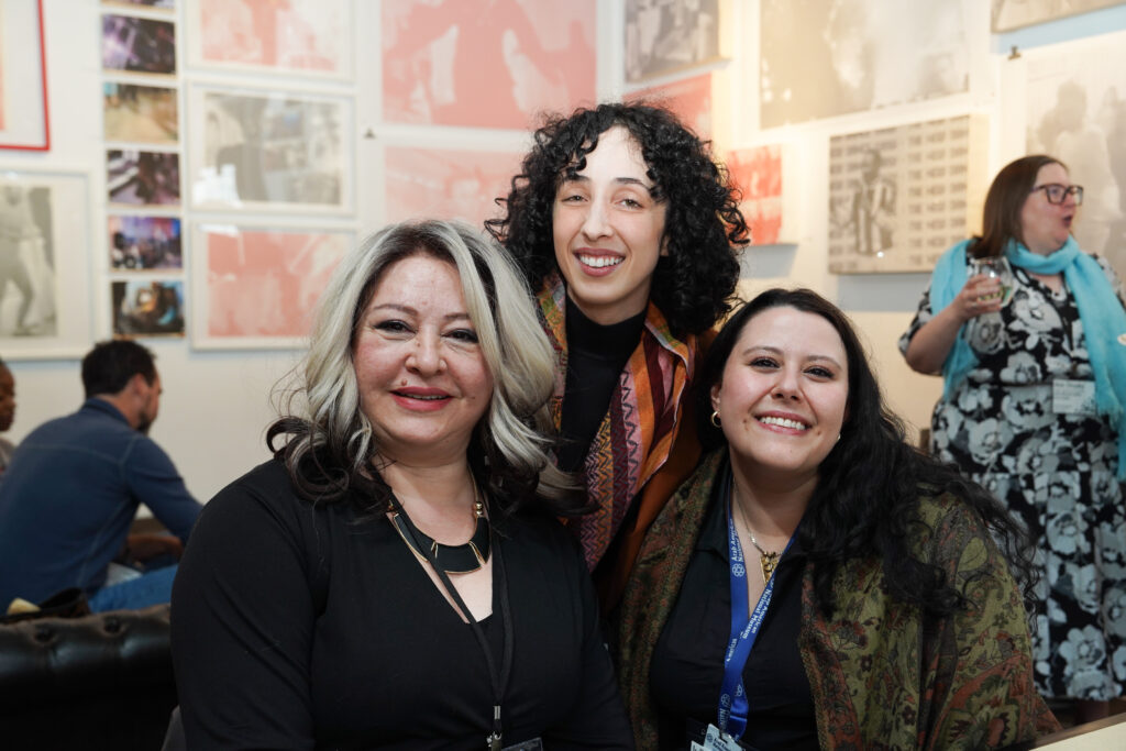 Three attendees of NPN's Community Reception at Spot Lite Detroit smile for a portrait. Behind them there are other attendees making conversation and artwork hung from the walls.