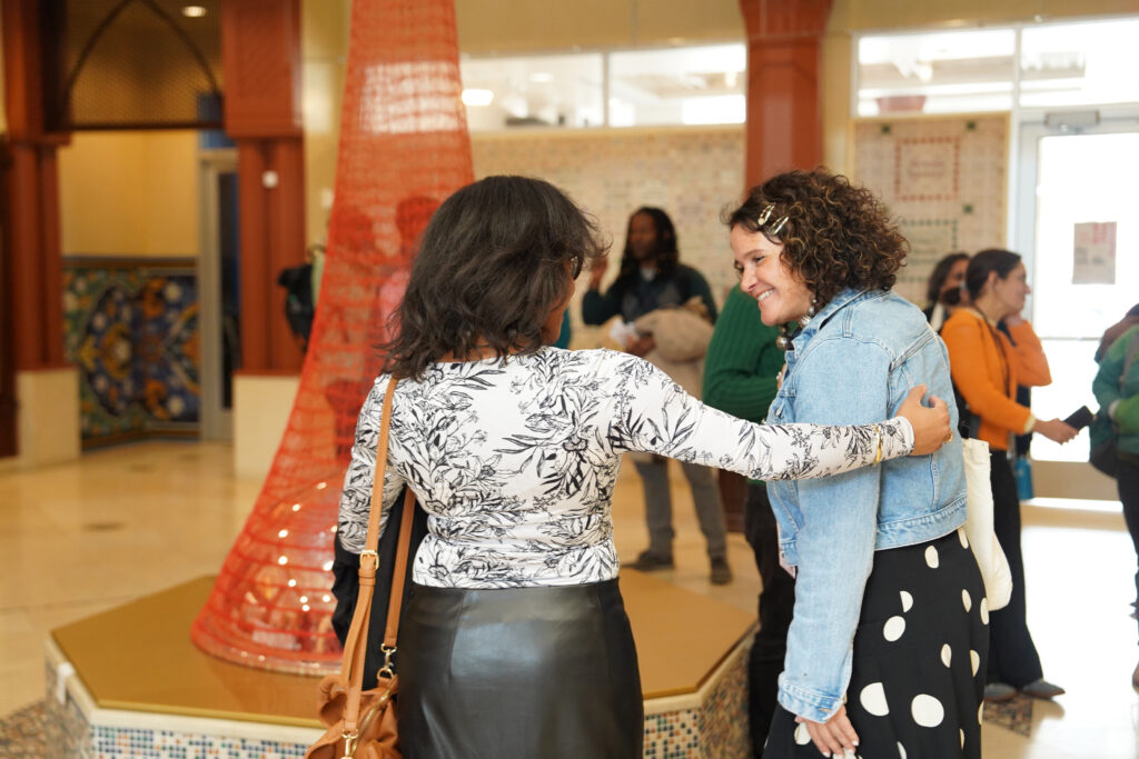 Two attendees of NPN's Partner Meeting greet each other in the Community Courtyard of the Arab American National Museum. In the background a sculptural work made of woven fabric rises from the center of the room and other attendees are gathered in conversation around it. Image by Paisley Sutton, Winnie Media.