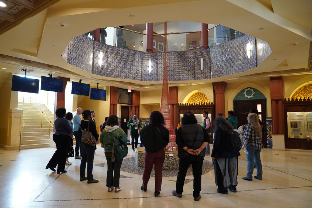 A small group of attendees from NPN's 2026 Partner Meeting in Dearborn gather in AANM's Community Courtyard for a tour of the museum. More than a dozen people gather around a sculptural work that rises up toward the museum's domed ceiling. 