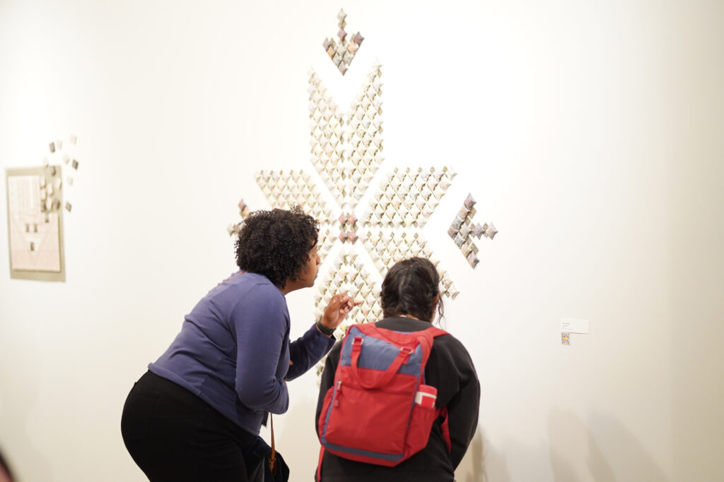 Two attendees of NPN's 2026 Partner Meeting in Dearborn crouch to inspect a porcelain tile artwork hung on the wall that is inspired by traditional Palestinian embroidery patterns.