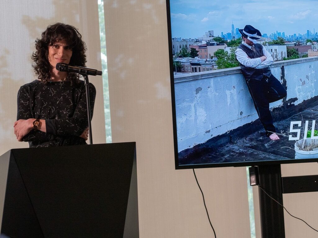 Poet Noa Micaela Fields stands behind a black podium. She is wearing a long-sleeve shirt with constellations printed on it. An image of the artist Nat Pyper is visible on a screen beside Noa. They are standing barefoot on a rooftop with a skyline visible in the background and are wearing a suit vest and a cowboy hat with a pair of paper dice affixed to the band of the hat.