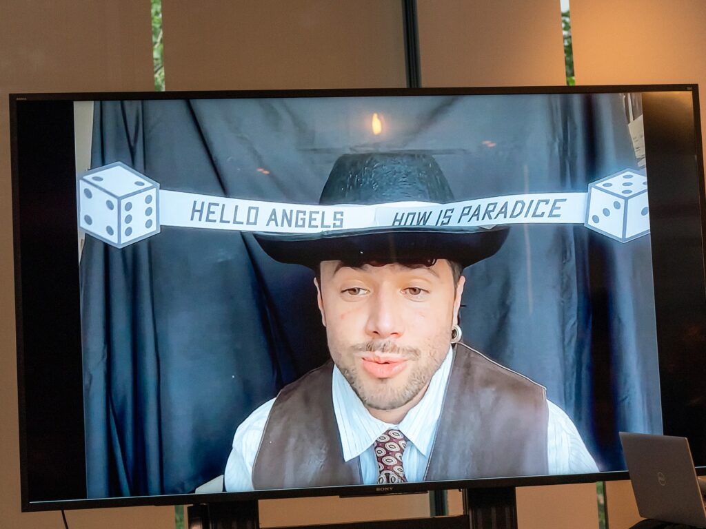Artist Nat Pyper speaks to the audience from a screen. They are seated in front of a black backdrop and are wearing a cowboy hat with two two-dimensional dice affixed to the band of the hat by a long strip of paper that reads, "Hello Angels How is Paradice." They are wearing a leather vest, a striped button-up shirt, and a tie. Photo by Larry Wolf.