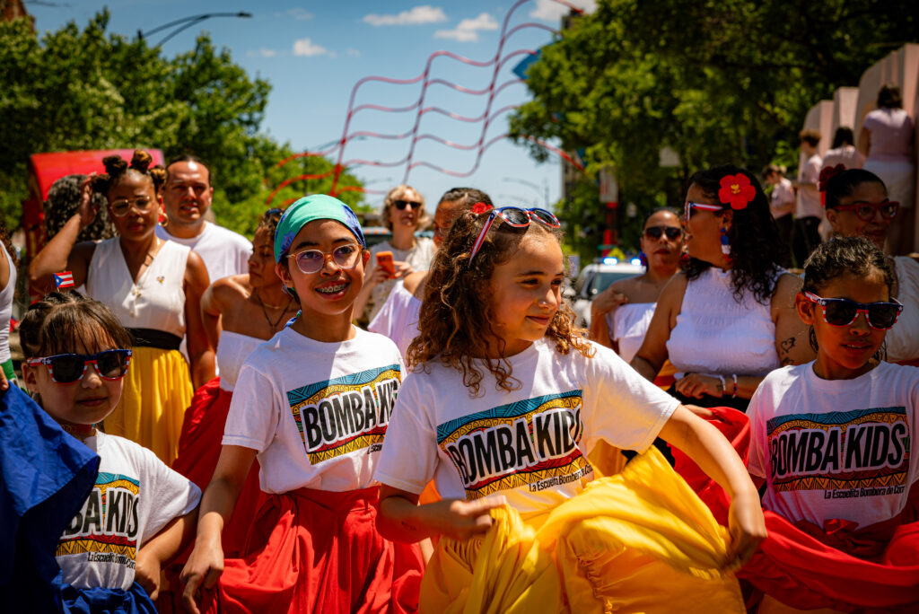 A group of kids from the La Escuelita Bombera de Corazón dance. In the background are more dancers and the large metal Puerto Rican flag. Photo by Tonal Mondae.