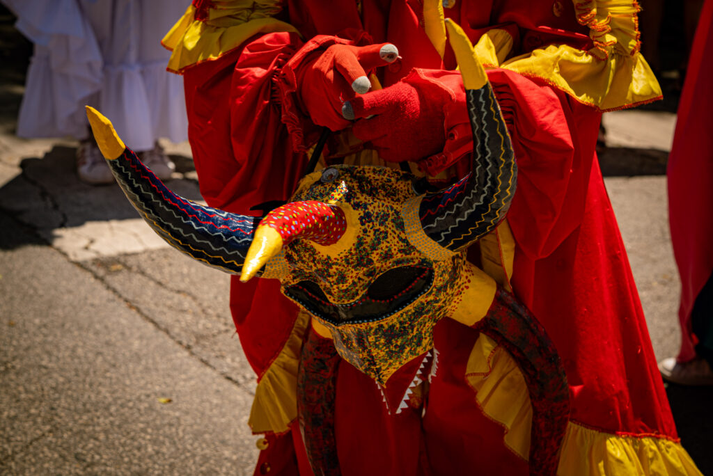 A close-up shot of a yellow, red, black, and white vejigante mask and the wearer holding it in their hands. It’s mostly yellow with designs on each of the five horns. Photo by Tonal Mondae. 