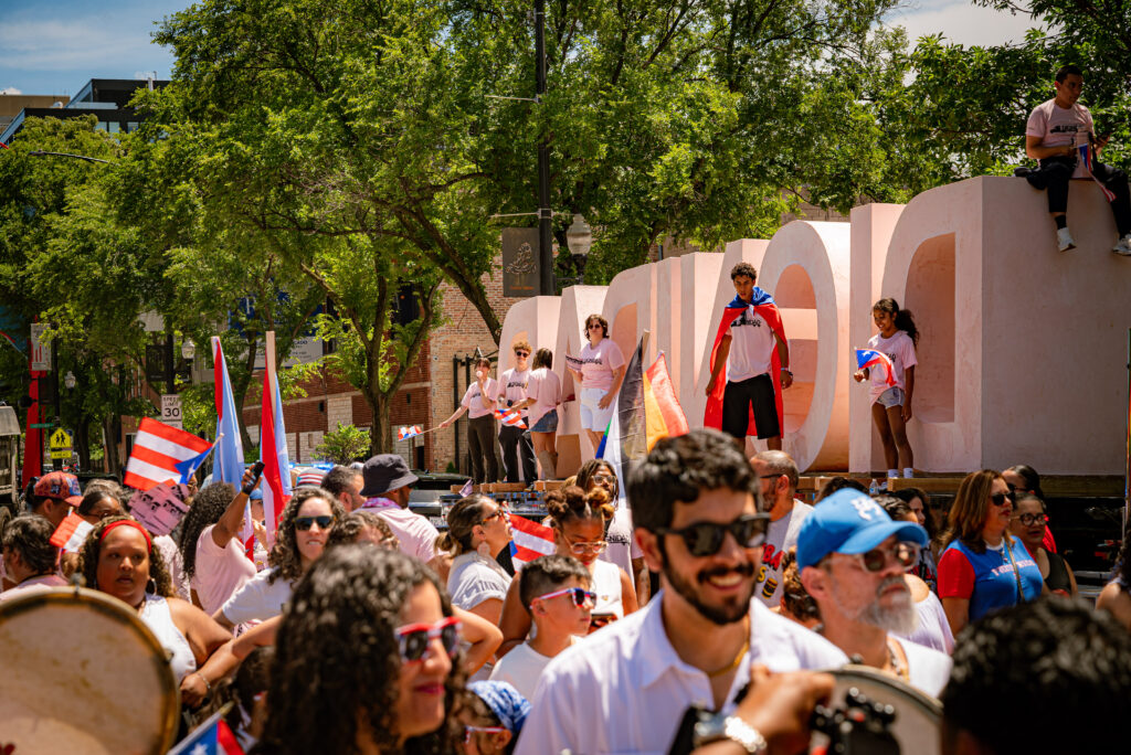 Participants of the Puerto Rican Day Parade stand in the street, and six people stand on top of the flatbed truck in front of the DIGNIDAD sculpture. Photo by Tonal Mondae.