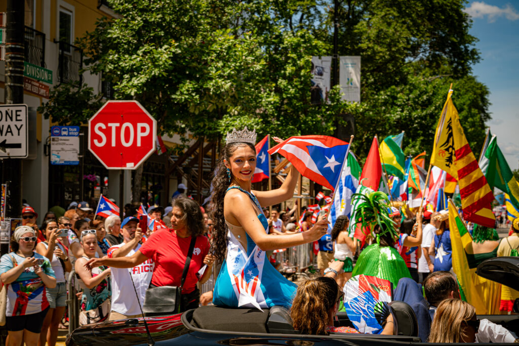 Miss Puerto Rico 2026 sits on top of a black convertible, waving a small handheld Puerto Rican flag at the camera in front of a crowd of people. Photos by Tonal Mondae.