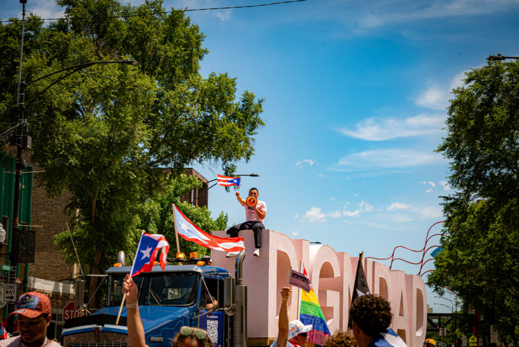 Artist Ivan Argoté sits atop the DIGNIDAD sculpture being led in the procession on a flatbed truck, waving the Puerto Rican flag and chanting “dignidad” from a microphone. Photo by Tonal Mondae.