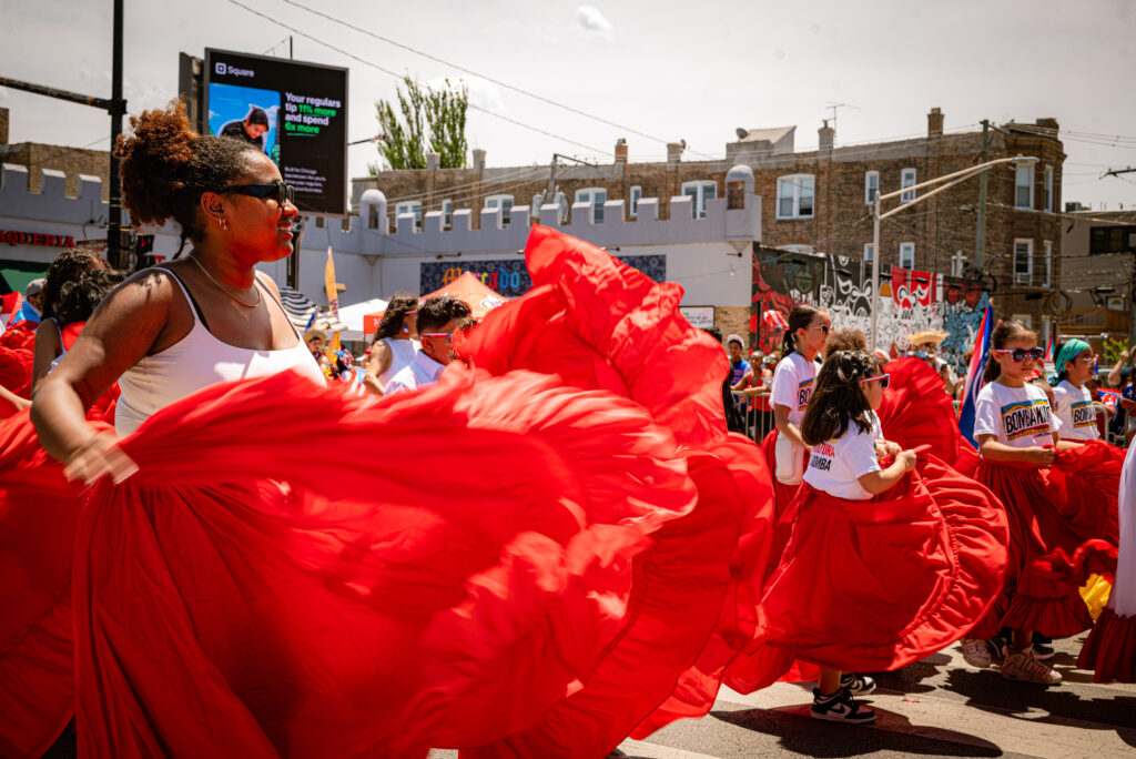 La Escuelita Bombera de Corazón dancers move with large red skirts down Division St in front of ¡WEPA! Mercado del Pueblo. Photo by Tonal Mondae.