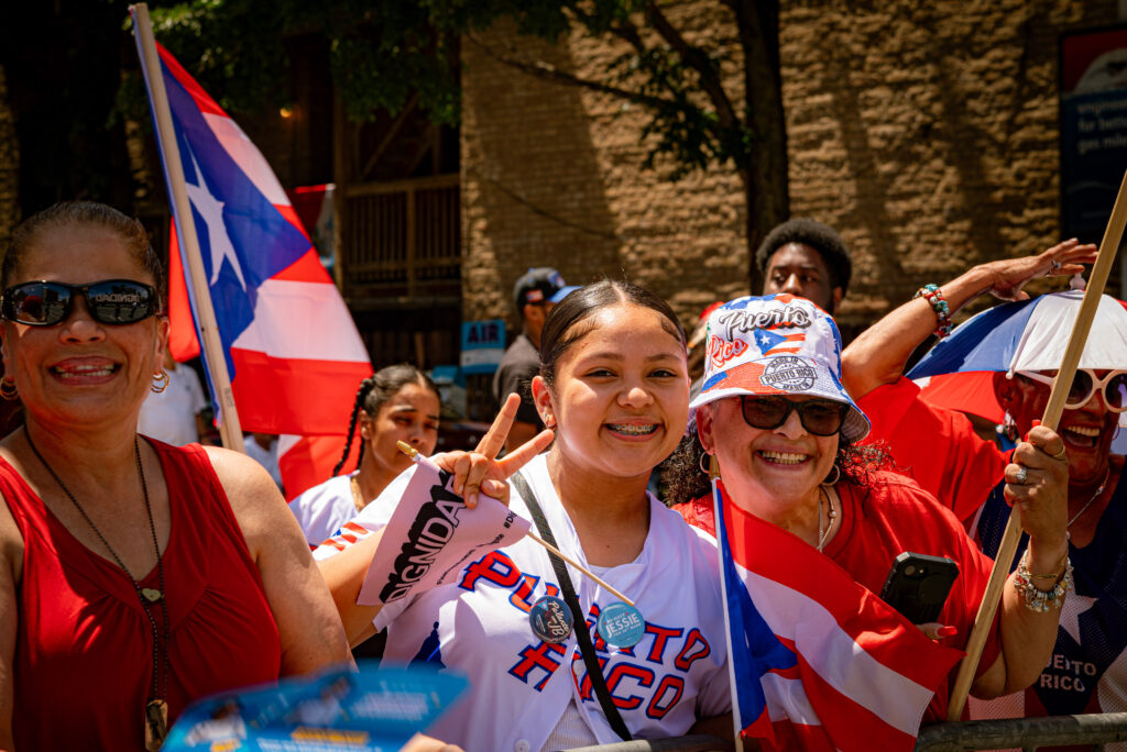 Crowd members wearing Puerto Rican colors and paraphernalia smile and look at the camera. Photo by Tonal Mondae.