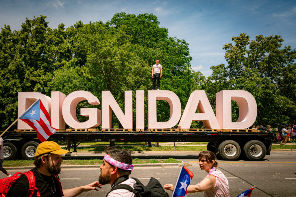 Artist Ivan Argoté stands atop the DIGNIDAD sculpture. The first ‘D’ is partially covered by the Puerto Rican flag, and he stands on the second ‘D.’ Photo by Tonal Mondae.