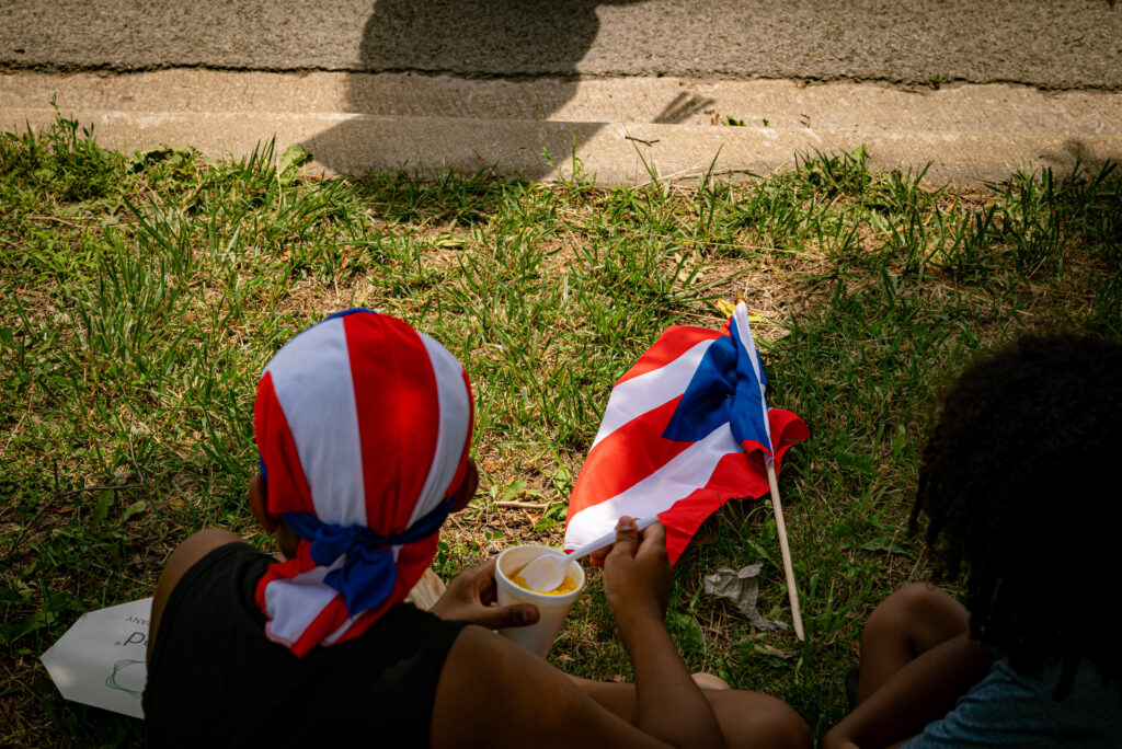 Two kids sit on the grass curb. One is eating frozen ice as they look onward toward the parade. In the middle of them lies the Puerto Rican flag on the ground. Photo by Tonal Mondae.