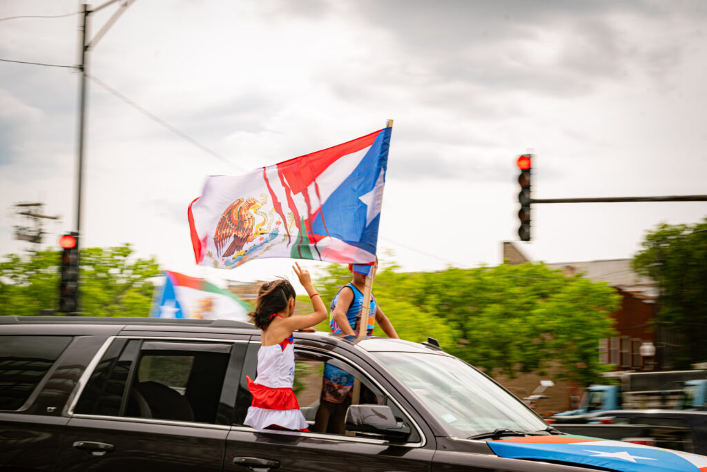 Two kids sit out the window of a black truck with a Mexican-Puerto Rican flag. One throws up the peace sign. Photo by Tonal Mondae. 
