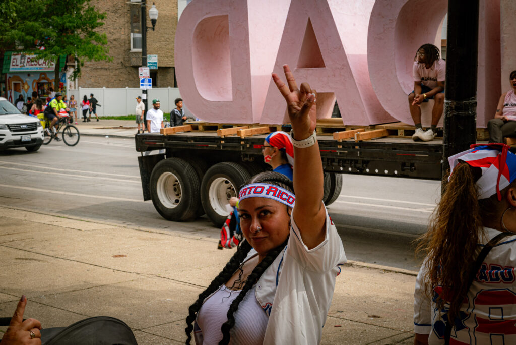 A person with a Puerto Rican headband throws up the peace sign looking into the camera. They are standing on the sidewalk between the Le Lo Lai Gallery and the parked DIGNIDAD sculpture. Photo by Tonal Mondae.