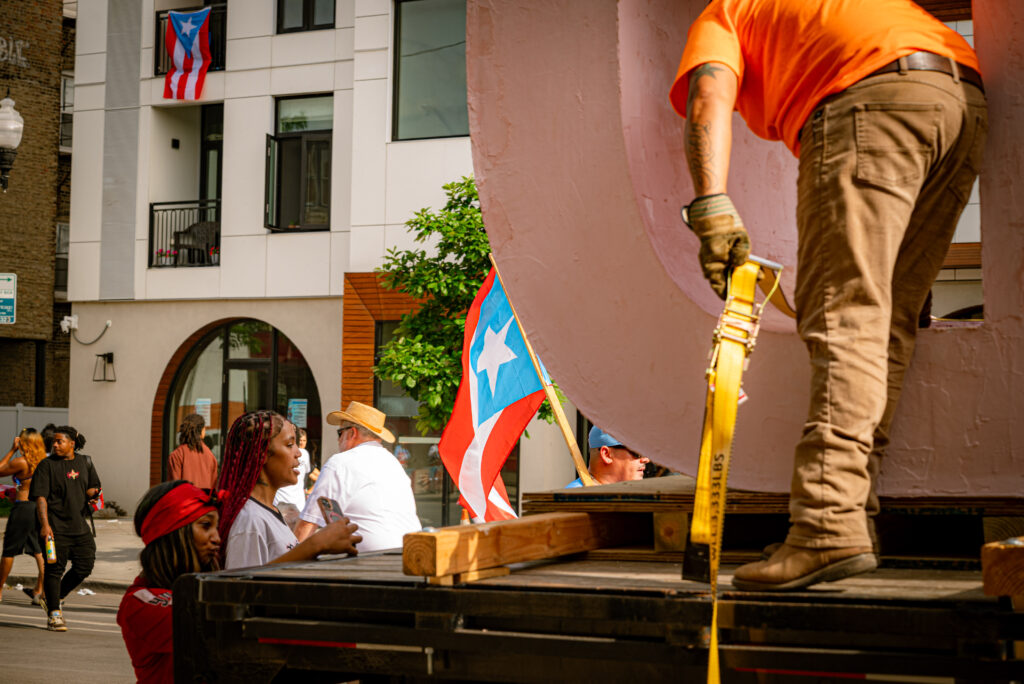 A truck driver is tying down the letters of the sculpture onto the flatbed for transport while another person uses the back of the truck to take a selfie. Photo by Tonal Mondae.