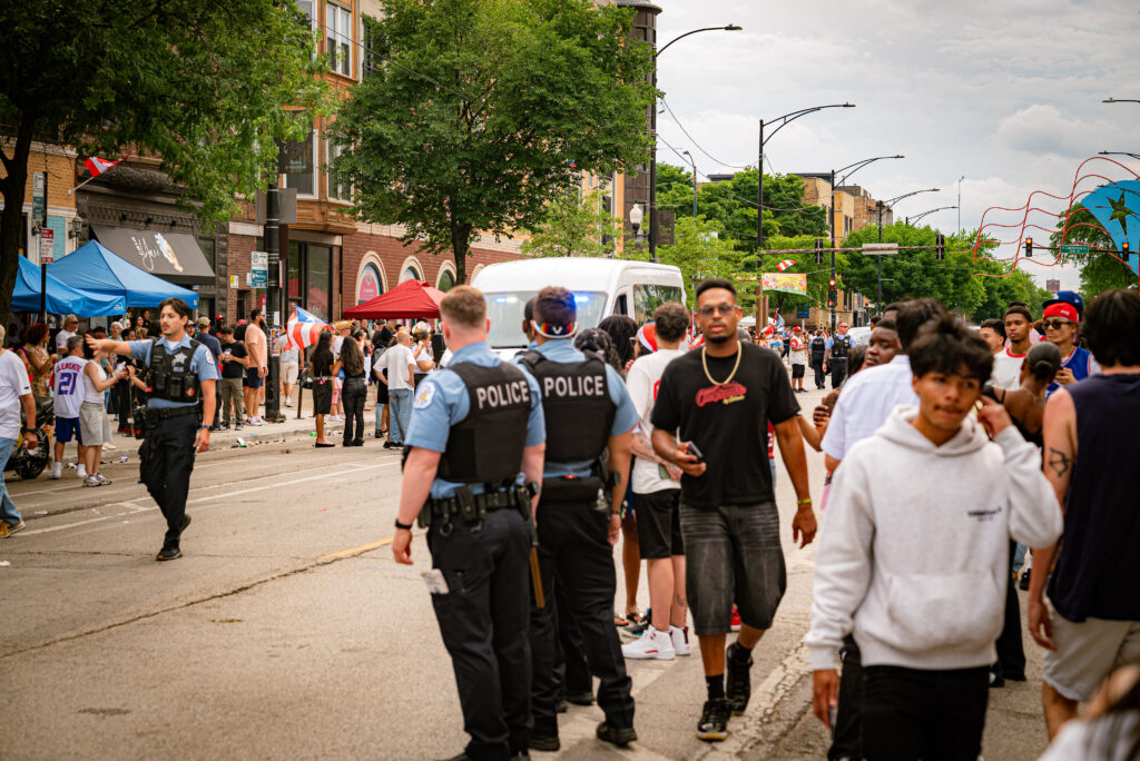 One police officer ushers the crowd to break up so a police van can drive through as two other officers stand to the side. Photo by Tonal Mondae.
