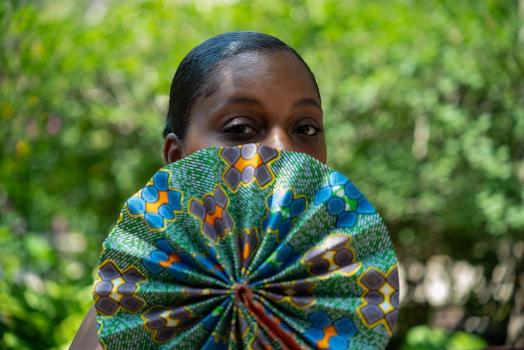 Rebelle Cunt looking over a multicolored patterned handheld fan with green nature background. Photo by Tonal Mondae.