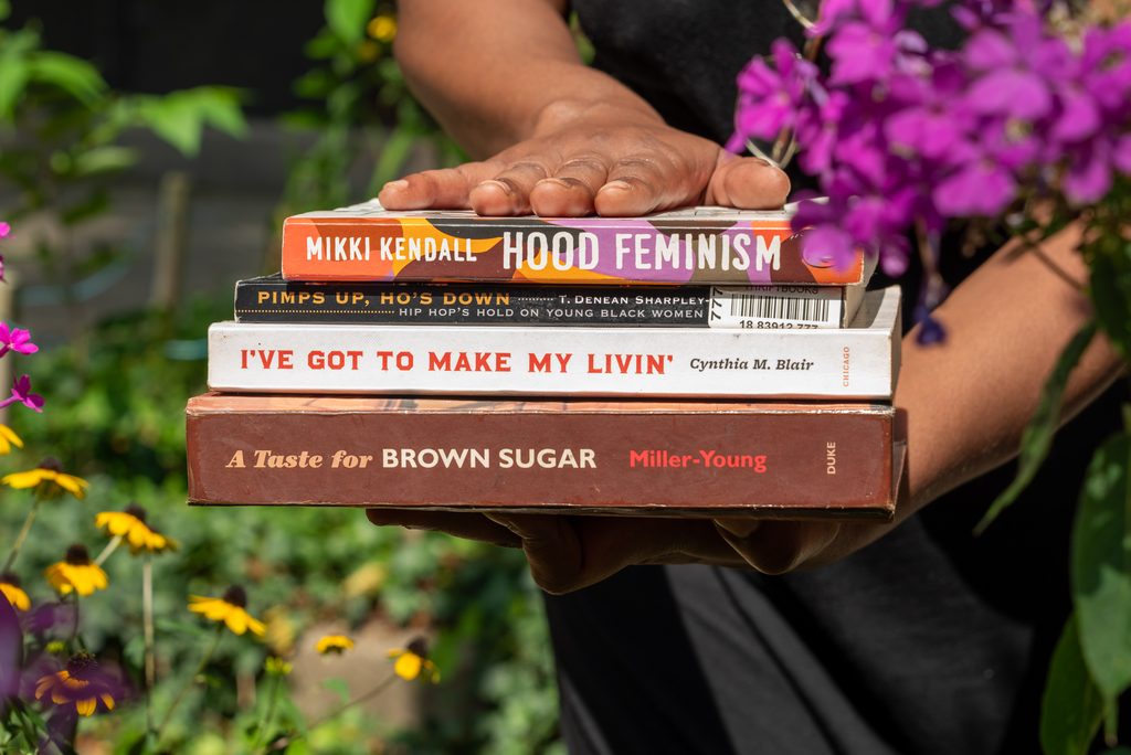 Rebelle Cunt pictured holding a stack of books: Hood Feminism by Mikki Kendall, Pimps Up, Ho's Down by T. Denean Sharpley, I've Got To Make My Livin' by Cynthia M. Blair, A Taste for Brown Sugar by Miller-Young. Photo by Tonal Mondae.