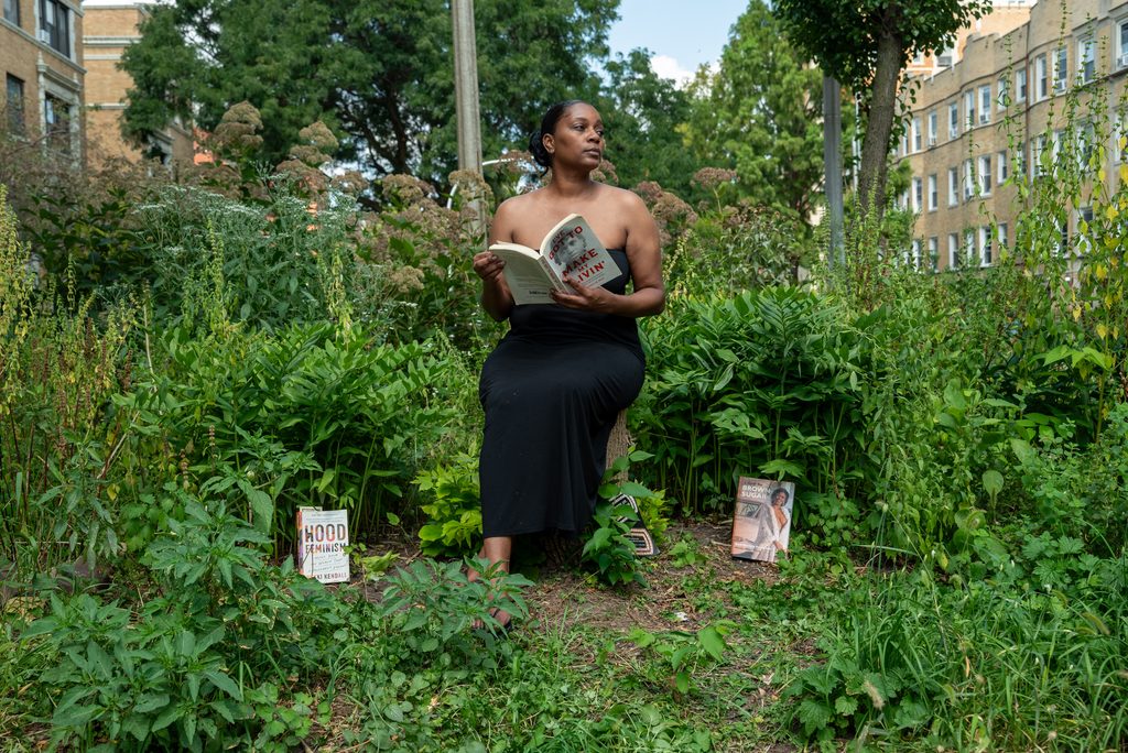 Rebelle Cunt captured reading a book while sitting in a garden surrounded by tall green plants with apartment buildings in the background. Photo by Tonal Mondae.