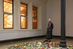 Adda Farcus within their exhibition Room Tone, Eureka College, 2026. Adda stands with their hands behind their back next to three large windows covered in orange lighting gels. At their feet are colorful flowers with green leaves, dark chunks of various sized rock-like fragments, and other found objects that are arranged in a large circle. Image courtesy of Adda Farcus. Photograph by Jade Minh-ha Nguyen.