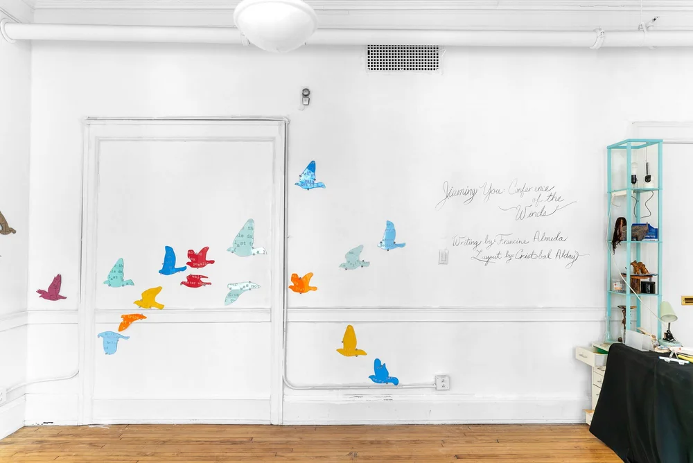 Installation capture of Conference of the Winds at HOOK Space. A group of colorful, silhouette-like starlings can be seen hung on the wall as if they are flying towards the left side of the gallery space. On the right there is writing on the wall that reads: "Jiaming You Conference of the Winds. Writing by Fraincine Almeda. Layout by Cristobal Alday." Photo Credit: Mikey Mosher.