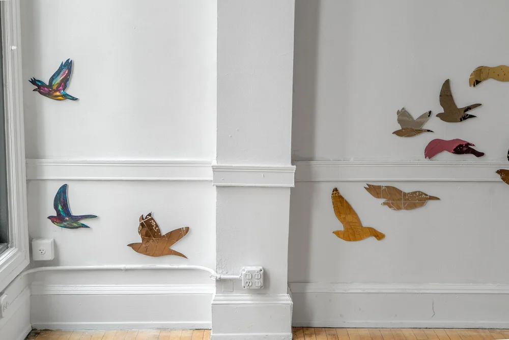 Installation capture of Conference of the Winds at HOOK Space. Various starling-shaped works can be seen flying across a white gallery wall. They are mostly brown and orange in color, with the two on the left being more colorful with shades of magenta, blue, green, and yellow. Photo Credit: Mikey Mosher.
