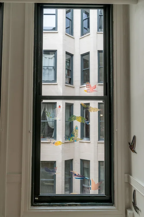 Installation image of Conference of the Winds at HOOK Space. A view of a window in the gallery space. Several translucent starlings can be seen displayed on the window. Photo Credit: Mikey Mosher.
