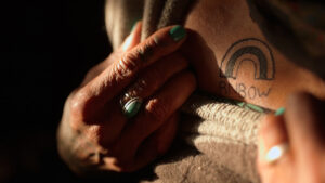 A close up on a pair of hands holding a rough, rope-type material. On the subject's skin is a black tattoo in the shape of a rainbow with the letters R N B O W beneath. The subjects green nails match the green hue of the ring on their finger. Photo by na forest lim.