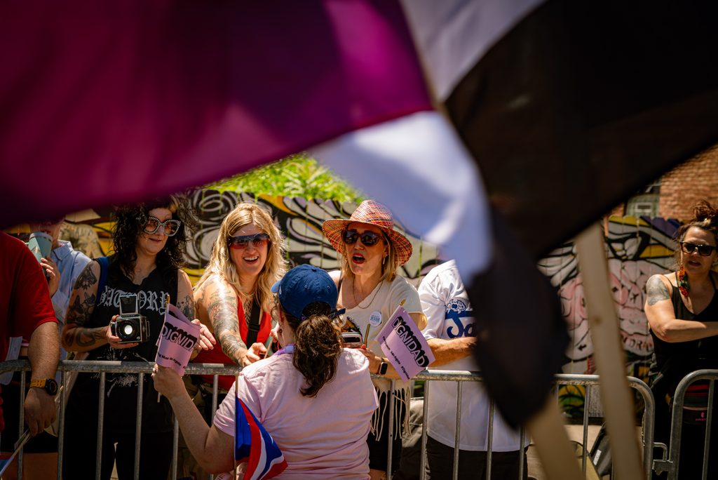Handheld pink flags bearing DIGNIDAD written in black are passed out to viewers behind the metal gate barricade. Photo by Tonal Mondae.