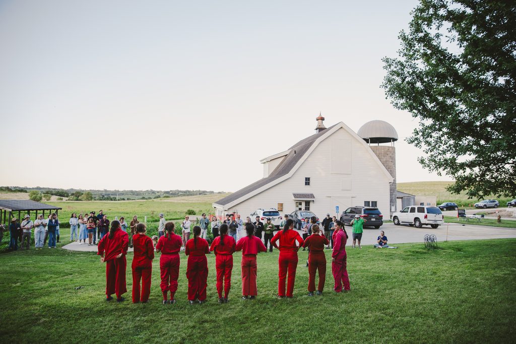tbd dance collective performers, all dressed up in red, line up in front of their audience on the mowed green grasses of Glacier Creek Preserve in front of its barn. The audience is pictured standing towards the parking lot of the barn in a line parallel to tbd dance collective performers. Image by Ben Semisch.