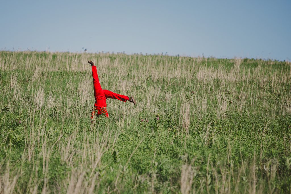 Shot from "Prairie Violet" choreographed by Kenzie Sawyer. Sawyer is pictured in red doing a headstand in the middle of the field prairie grasses at Glacier Creek Preserve. Image by Ben Semisch.