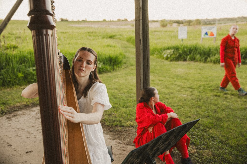Elyse N. Jorgensen, dressed in a white dress, plays the harp with a music stand in front of her, on the grounds of Glacier Creek Preserve during a performance of "Passages" by tbd dance collective. A performer is pictured seated on the ground behind her while another performer dressed in red is pictured walking past the other performer. The performers in red are staring directly at each other. Image by Ben Semisch.