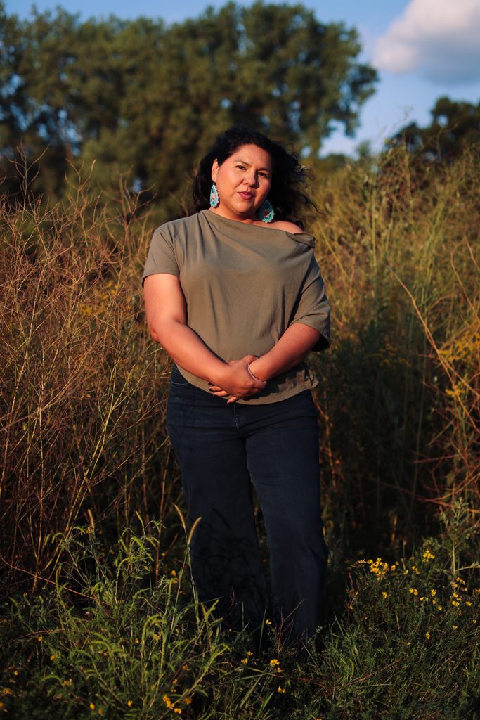 Jaida Grey Eagle captured by Patience Zalanga. She is wearing a green off shoulder t-shirt while smirking and holding her hands. She is standing in a field with tall greenery around her.