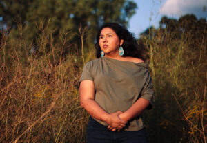 Jaida Grey Eagle captured by Patience Zalanga. She is standing in a field with tall greenery around her while wearing a green off shoulder t-shirt and looking away from the camera.