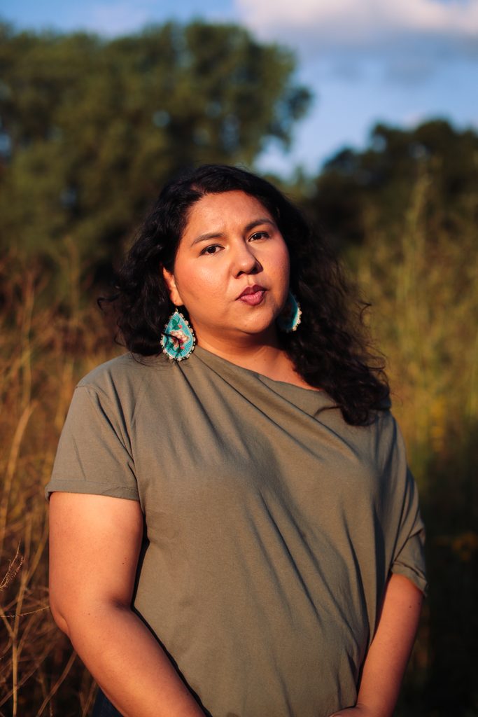 Jaida Grey Eagle captured by Patience Zalanga. She is wearing a green t-shirt and teal beaded earrings with an image of a hummingbird above a flower. 