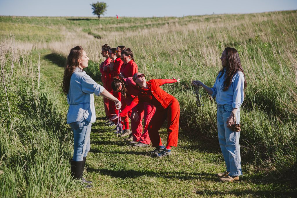 Shot from a performance of "Butterfly Milkweed" choreographed by Gayle Rocz with original music composed & performed by Pearl Lovejoy Boyd and Susan Sanchez Olson, who are dressed in blue and standing closest to the camera. The rest of tbd dance collective formed a line down the center of a walking path on the prairie and in between the Pearl Lovejoy Boyd and Susan Sanchez who are facing each other. The tall grasses surround the performers. Image by Ben Semisch.