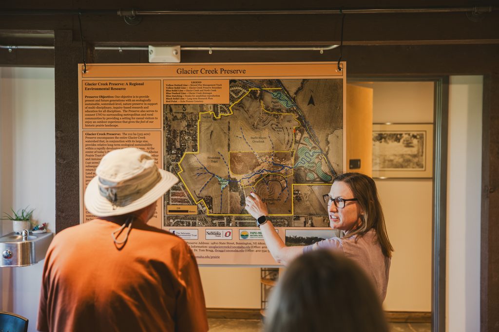 Glacier Creek Preserve tour guide wearing an Apple watch, black-rimmed glasses and a mauve t-shirt, points at a map of the creek while speaking to a tour group inside of an informational building and rest stop on a the preserve. The tour guide's gaze is focused on a tour participant who is wearing an orange long-sleeved shirt and bucket hat. Image by Ben Semisch.
