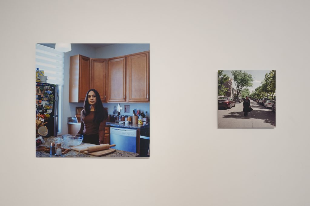 Image: Two unframed portraits hang on a white wall. The photo on the left depicts a person in a kitchen. Behind them are brown cabinets and a dishwasher. In front of them is a counter with a mixing bowl, rolling pin, and cutting board. The image on the right depicts a person clad in dark colors standing outside, in the middle of a car-lined street on a sunny day. Image courtesy of Columbia College Chicago. Photo by Alex Stahr.
