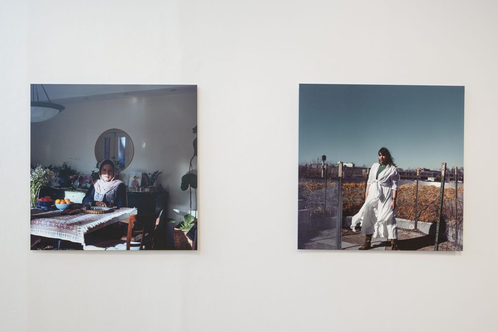Image: Two unframed portraits hang on a white wall. The photo on the left depicts a person wearing a headscarf and sitting at a dining table. Sunlight streams in, but part of their face is partially cast in shadow. The image on the right depicts a person standing outside, wearing a white gown that billows slightly in the wind. Image courtesy of Columbia College Chicago. Photo by Alex Stahr.