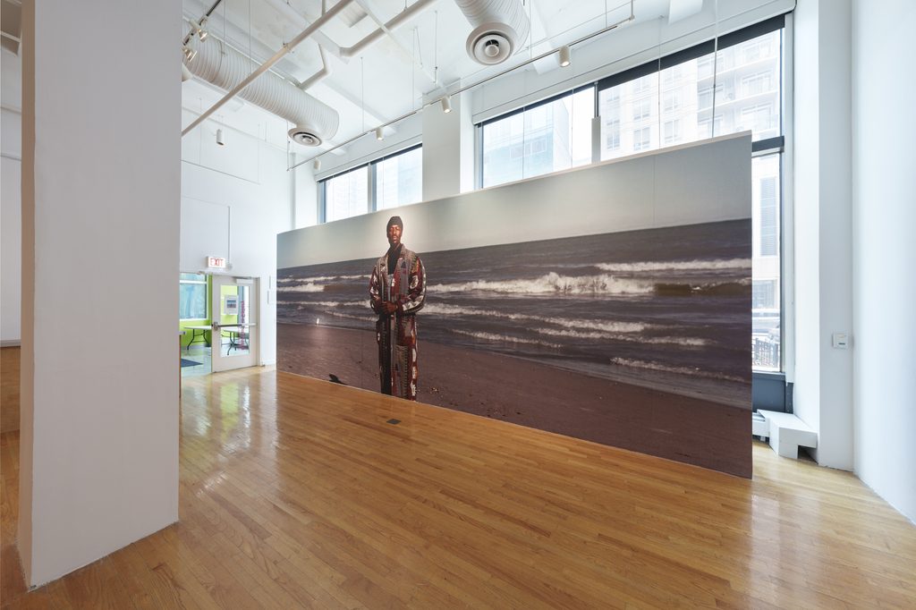 Image: Installation view of The Distance We Carry. A freestanding photograph blown up to span nearly the entire room. In the image, a person in a long coat and winter hat stands on a beach in front of a large body of water. The waves break behind them beneath a blue-gray sky. Image courtesy of Columbia College Chicago. Photo by Alex Stahr.