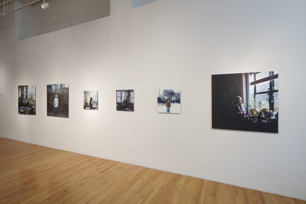 Image: Installation photo of The Distance We Carry. Six unframed portraits in various sizes hang on a white wall. Each image depicts a person in a different setting. Some are photographed outside; some are photographed inside. All of the images are unique. Image courtesy of Columbia College Chicago. Photo by Alex Stahr.