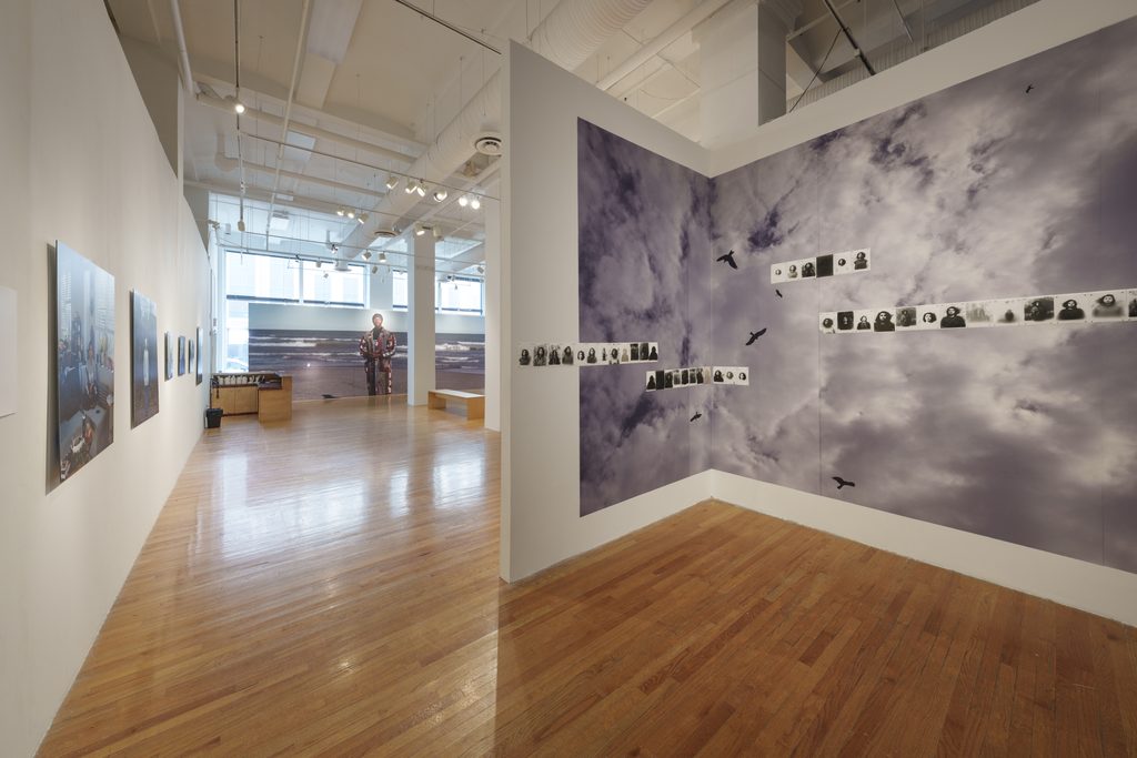 Image: Installation view of The Distance We Carry. Installed on the L-shaped wall is a large-scale black-and-white image of the sky with fluffy white clouds and black birds circling in the air. Offset over the image are four rows of black-and-white portraits. Image courtesy of Columbia College Chicago. Photo by Alex Stahr.