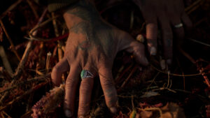 Image: A closeup on a pair of hands digging in the earth. The fingertips clutch branches, dirt, and other natural materials. A warm light illuminates one hand with a green ring, while the other hand is hidden in shadows cast by the light. Photo by na forest lim.