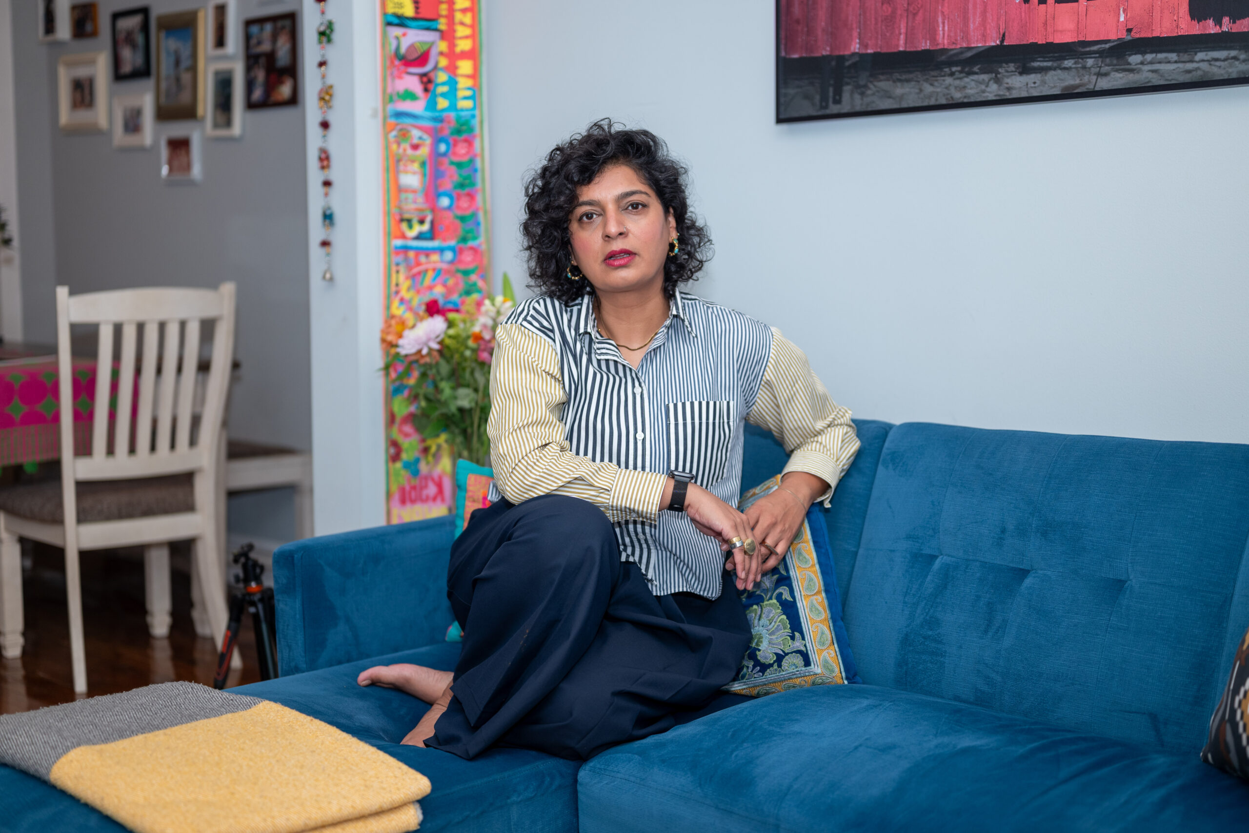 Image: The artist Mehreen Lali Khalid in her colorfully decorated home. Sitting on a blue couch with her legs folded underneath her, she's clad in a striped blouse and facing the viewer. Behind her, out of focus, is a bouquet of flowers. Photo by Tonal Mondae.