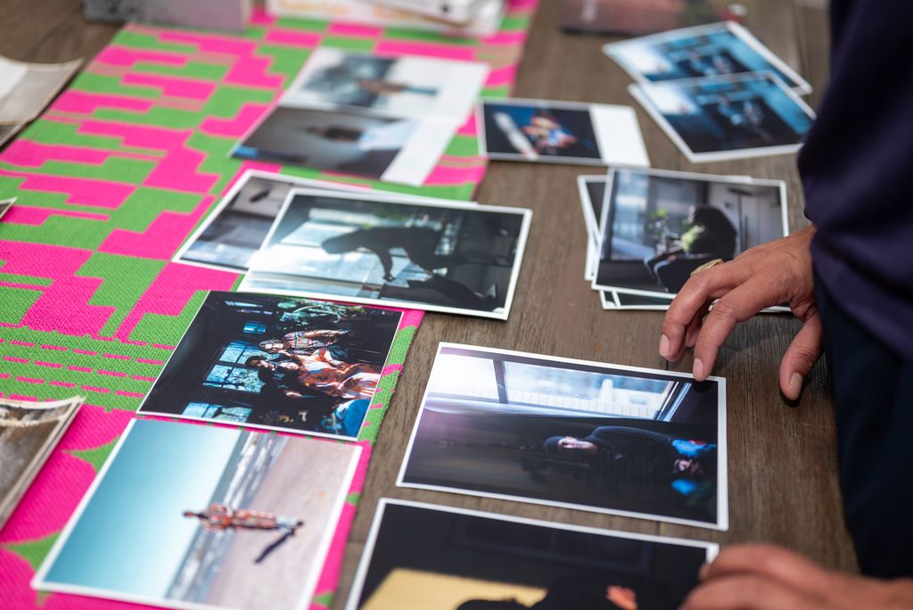 Image: A few photos from the exhibition lay unframed on a wooden table. Some images are organized in small piles; others lie atop a table runner with a pink and green geometric design. The artist's hands are visible in the image; one hand gently touches the corner of a photo. Photo by Tonal Mondae.