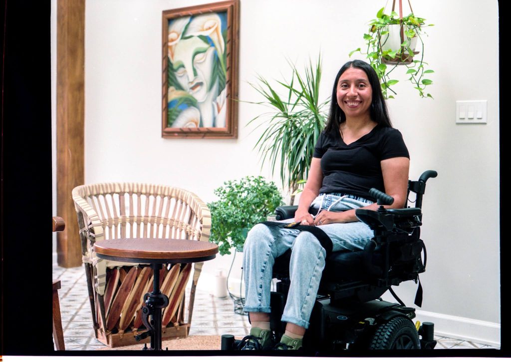 A photo of Reveca sitting in her motorized wheelchair, her straight brown hair is down and parted slightly to the side. She is smiling and wearing a black V-neck T-shirt and blue jeans. Next to her is a low, round wooden table and gorgeous woven and wooden chair with warm, variegated, diagonal wood slats on the bottom and a lighter wood colored lattice strung around the back. The walls are painted a bright, clean, white and there are small, patterned, square tiles lining the floor. Reveca is framed from the left by three healthy plants, one of them draping down behind her from a hanging basket. Further to the left of her face is a surrealist portrait of a woman's face with green and blue shadowing to highlight the contours of the face and orange calla lilies around her head in place of hair. Photograph by Genesis Falls.