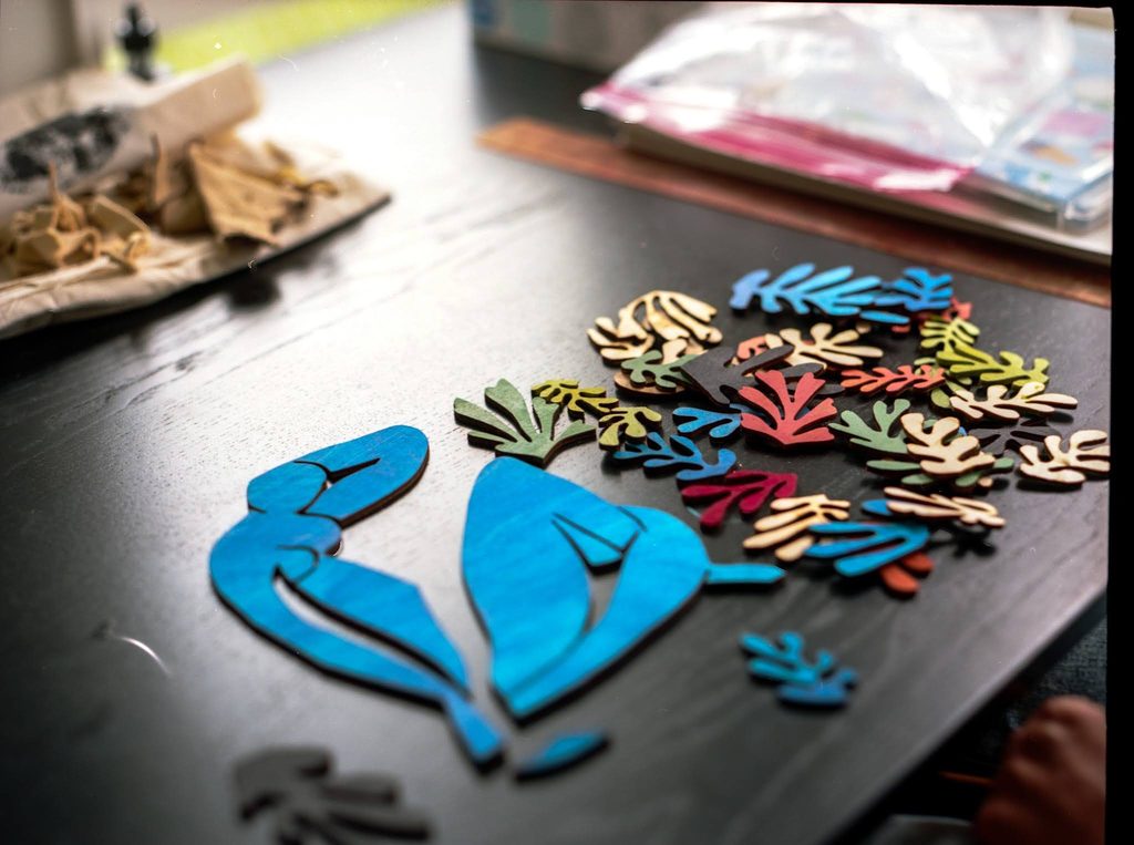 Deconstructed pieces of a Matisse work called le gerbe made out of laser cut wood that has been painted in bright colors. There is a blue representation of a seated figure of a woman's body next to a pile of individually cut out, different colored leaves. The pieces are on a black wooden table next to the corner of a pile of mustard colored fabric scraps. Photograph by Genesis Falls.