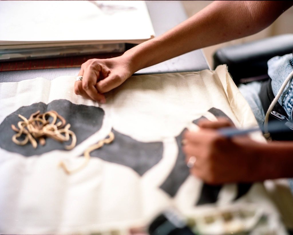A photo from above of Reveca’s arms from the elbow down resting on a table. She is working on a painting. One hand in the background is gently curved and resting on a canvas which has big black shapes painted on it, and a little pile of leather cord. A blurry hand in the foreground is holding a paintbrush and adding black to one of the curved shapes. Photograph by Genesis Falls.